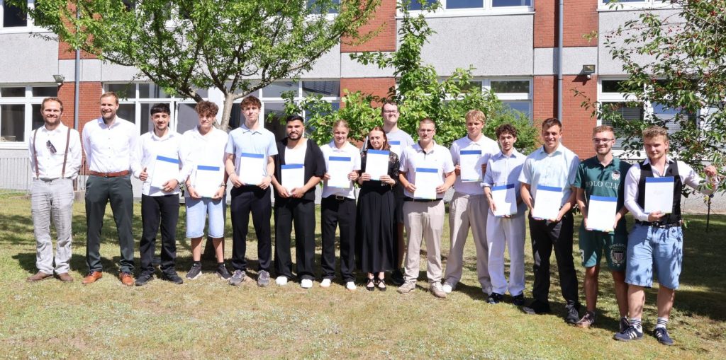 Eine Gruppe von Jugendlichen und Erwachsenen steht im Halbkreis auf einer Rasenfläche vor einem Schulgebäude. Die Jugendlichen tragen überwiegend helle, sommerliche Kleidung und halten jeweils eine Urkunde in den Händen. Zwei Erwachsene stehen am linken Bildrand, beide in Hemd und Hose, einer davon mit Krawatte. Im Hintergrund sind Bäume und das mehrstöckige Gebäude mit großen Fenstern und roten Klinkerelementen zu sehen. Die Szene ist von der Abschlussfeier der Berufsfachschule Technik 2026.