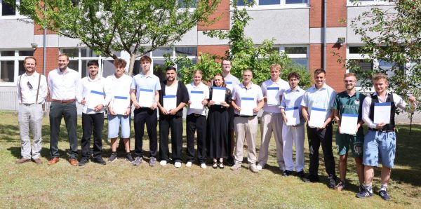 Eine Gruppe von Jugendlichen und Erwachsenen steht im Halbkreis auf einer Rasenfläche vor einem Schulgebäude. Die Jugendlichen tragen überwiegend helle, sommerliche Kleidung und halten jeweils eine Urkunde in den Händen. Zwei Erwachsene stehen am linken Bildrand, beide in Hemd und Hose, einer davon mit Krawatte. Im Hintergrund sind Bäume und das mehrstöckige Gebäude mit großen Fenstern und roten Klinkerelementen zu sehen. Die Szene ist von der Abschlussfeier der Berufsfachschule Technik 2026.