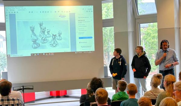 Im Vordergrund sitzen mehrere Schülerinnen und Schüler in einem hellen Raum und blicken auf eine große Leinwand. Drei Jugendliche und ein Erwachsener stehen seitlich vor der Leinwand. Der Erwachsene hält ein Mikrofon und spricht, während die Jugendlichen aufmerksam zuhören. Auf der Leinwand ist eine technische Zeichnung oder ein 3D-Modell von Schachfiguren zu sehen, die auf einem Computerbildschirm angezeigt wird. Die Szene vermittelt den Eindruck einer Präsentation oder eines Workshops im Rahmen des Enrichment-Programms des Landes Schleswig-Holstein, bei dem besonders begabte und interessierte Schülerinnen und Schüler gefördert werden.