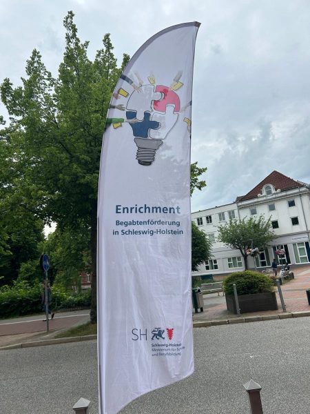 Im Vordergrund steht ein weißes, hoch aufragendes Banner mit dem Aufdruck des Enrichment-Programms zur Begabtenförderung in Schleswig-Holstein. Im oberen Bereich des Banners ist eine stilisierte Glühbirne abgebildet, deren Glaskolben aus vier Puzzleteilen besteht. Um die Glühbirne herum sind bunte Symbole angeordnet, die verschiedene Fachdisziplinen und Interessenfelder symbolisieren. Darunter steht in blauer Schrift: 'Enrichment Begabtenförderung in Schleswig-Holstein'. Im unteren Bereich befinden sich das Logo des Landes Schleswig-Holstein und ein weiteres, rotes Symbol. Im Hintergrund sind ein Baum, ein Gehweg, einige Poller und ein mehrstöckiges Gebäude mit rotem Dach zu sehen. Die Szene spielt im Freien bei bewölktem Himmel.