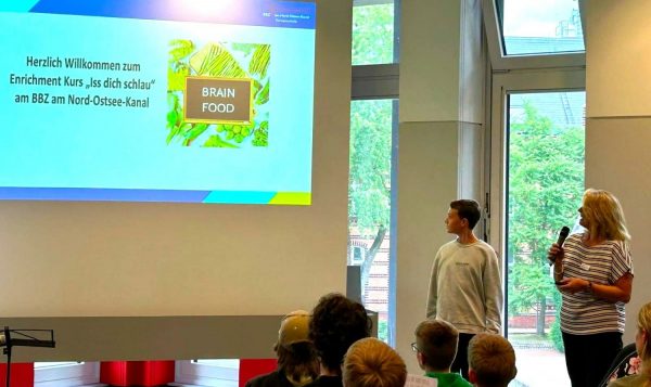 Eine Frau und ein Junge stehen nebeneinander vor einer Leinwand und blicken auf die Projektion. Die Frau hält ein Mikrofon. Auf der Leinwand ist eine Präsentationsfolie mit dem Titel 'Herzlich Willkommen zum Enrichment Kurs „Iss dich schlau“ am BBZ am Nord-Ostsee-Kanal' zu sehen. Daneben befindet sich ein Bild mit der Aufschrift 'BRAIN FOOD' auf grünem Hintergrund. Im Vordergrund sitzen mehrere Kinder und Jugendliche und schauen zur Bühne. Durch große Fenster fällt Tageslicht in den Raum, draußen sind Bäume und ein Backsteingebäude sichtbar. Die Szene spielt im Rahmen des Enrichment-Programms des Landes Schleswig-Holstein.