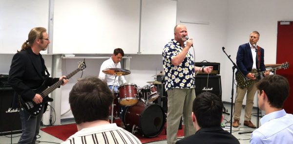 Eine Band spielt in einem Klassenraum . Im Vordergrund stehen mehrere Personen mit dem Rücken zur Kamera. Im Zentrum steht ein Sänger mit Glatze und Bart, der ein Mikrofon hält und ein kurzärmeliges Hemd mit Blumenmuster trägt. Links spielt ein Musiker E-Bass, rechts ein weiterer Musiker E-Gitarre. Im Hintergrund sitzt ein Schlagzeuger am Schlagzeug. Die Musiker sind von Verstärkern und Instrumenten umgeben. Die Szene ist aus einem Auftritt im Rahmen einer Abschlussfeier am BBZ am NOK.