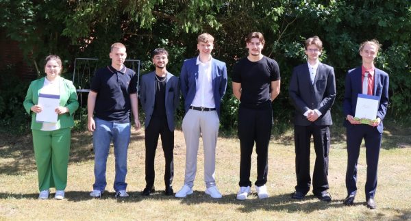 Eine Gruppe von jungen Erwachsenen steht nebeneinander auf einer Wiese vor einer Kulisse aus grünen Bäumen. Zwei Personen halten jeweils ein Dokument in der Hand, das auf eine feierliche Verabschiedung hinweist. Die Kleidung reicht von formellen Anzügen bis zu einem grünen Hosenanzug. Die Personen blicken in Richtung Kamera und lächeln. Das Bild vermittelt den Eindruck einer Abschlussfeier für die Technischen Assistenten des BBZ am NOK im Jahrgang 2026.