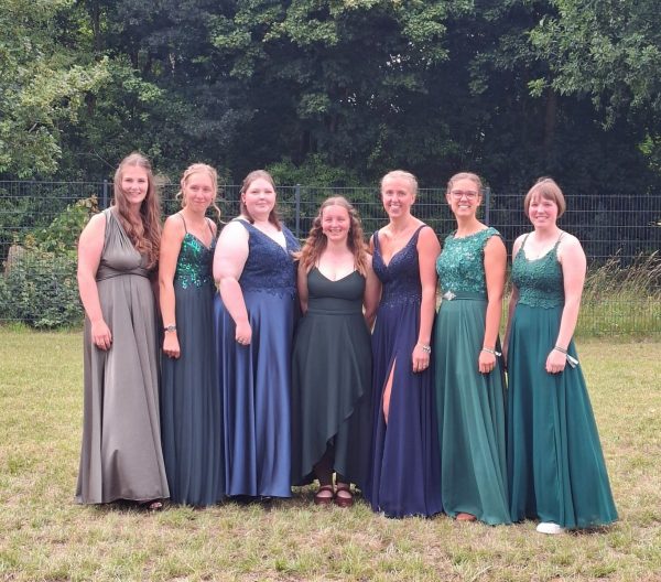 Sieben junge Frauen stehen nebeneinander auf einer Wiese vor einem Hintergrund aus Bäumen und einem Metallzaun. Sie tragen festliche, bodenlange Kleider in verschiedenen Grüntönen und Dunkelblau, einige mit Glitzerelementen. Die Frauen lächeln und blicken direkt in die Kamera. Die Szene zeigt den Abschlussjahrgang für Bildungsgangs zur hauswirtschaftlichen Betriebsleiterin am Standort Osterrönfeld des BBZ-NOK.
