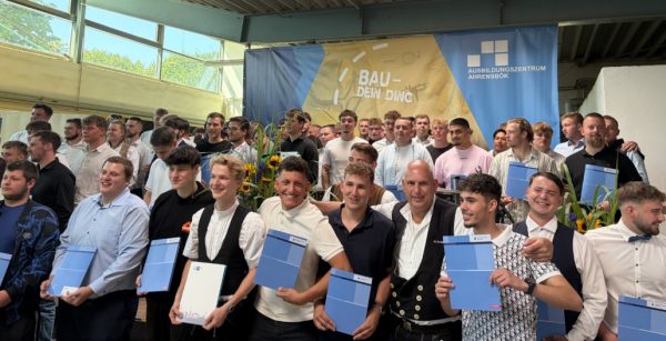 Im Vordergrund stehen mehrere junge Menschen, die blaue Urkunden oder Zertifikate in den Händen halten und lächeln. Sie tragen unterschiedliche festliche Kleidung, darunter Hemden, Anzüge und traditionelle Zunftkleidung. Im Hintergrund sind weitere Personen zu sehen, die eng beieinander stehen. Die Szenerie spielt in einer großen Halle mit Tageslicht, dekoriert mit Sonnenblumen und einem blauen Banner, auf dem das Logo des Ausbildungszentrums Ahrensbök sowie der Schriftzug 'BAU – DEIN DING' zu erkennen sind. Die Atmosphäre wirkt feierlich und gemeinschaftlich.