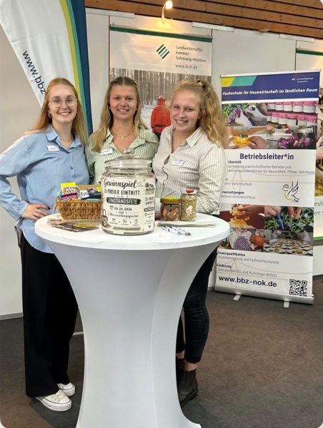 Im Vordergrund steht ein runder, weißer Stehtisch, auf dem ein dekorativer Behälter mit der Aufschrift 'Gewinnspiel' sowie zwei Körbe mit verschiedenen Produkten arrangiert sind. Dahinter stehen drei junge Frauen, die lächeln und Namensschilder tragen. Im Hintergrund sind mehrere Roll-Up-Banner zu sehen, darunter eines mit dem Logo des BBZ am Nord-Ostsee-Kanal und eines mit dem Schriftzug 'Betriebsleiter*in', das auf die Fachrichtung Hauswirtschaft im ländlichen Raum hinweist. Die Szene zeigt einen Messestand des BBZ am Nord-Ostsee-Kanal auf der NORLA, betreut von Schülerinnen und Lehrkräften aus den Bereichen Hauswirtschaft und Landwirtschaft.