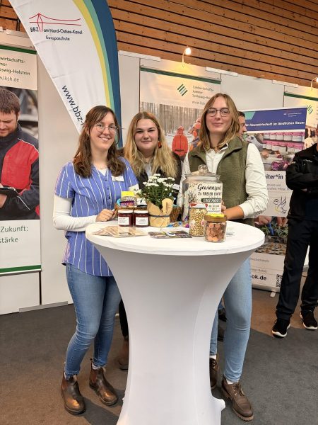 Im Vordergrund steht ein runder, weißer Stehtisch, auf dem verschiedene Lebensmittel wie Marmeladengläser, ein Glas mit eingelegtem Gemüse, ein kleiner Blumenstrauß und weitere Produkte arrangiert sind. Drei junge Frauen stehen hinter dem Tisch und lächeln in Richtung Kamera. Sie tragen legere Kleidung, darunter Jeans und Oberteile in unterschiedlichen Farben. Im Hintergrund sind mehrere Roll-Up-Banner mit dem Logo und Schriftzug des BBZ am Nord-Ostsee-Kanal sowie weitere Informationsmaterialien zu sehen. Die Szenerie befindet sich in einer Messehalle mit Holzverkleidung an der Wand. Das Bild vermittelt den Eindruck eines Messestandes, der von Schülerinnen und Lehrkräften aus den Bereichen Hauswirtschaft und Landwirtschaft betreut wird.